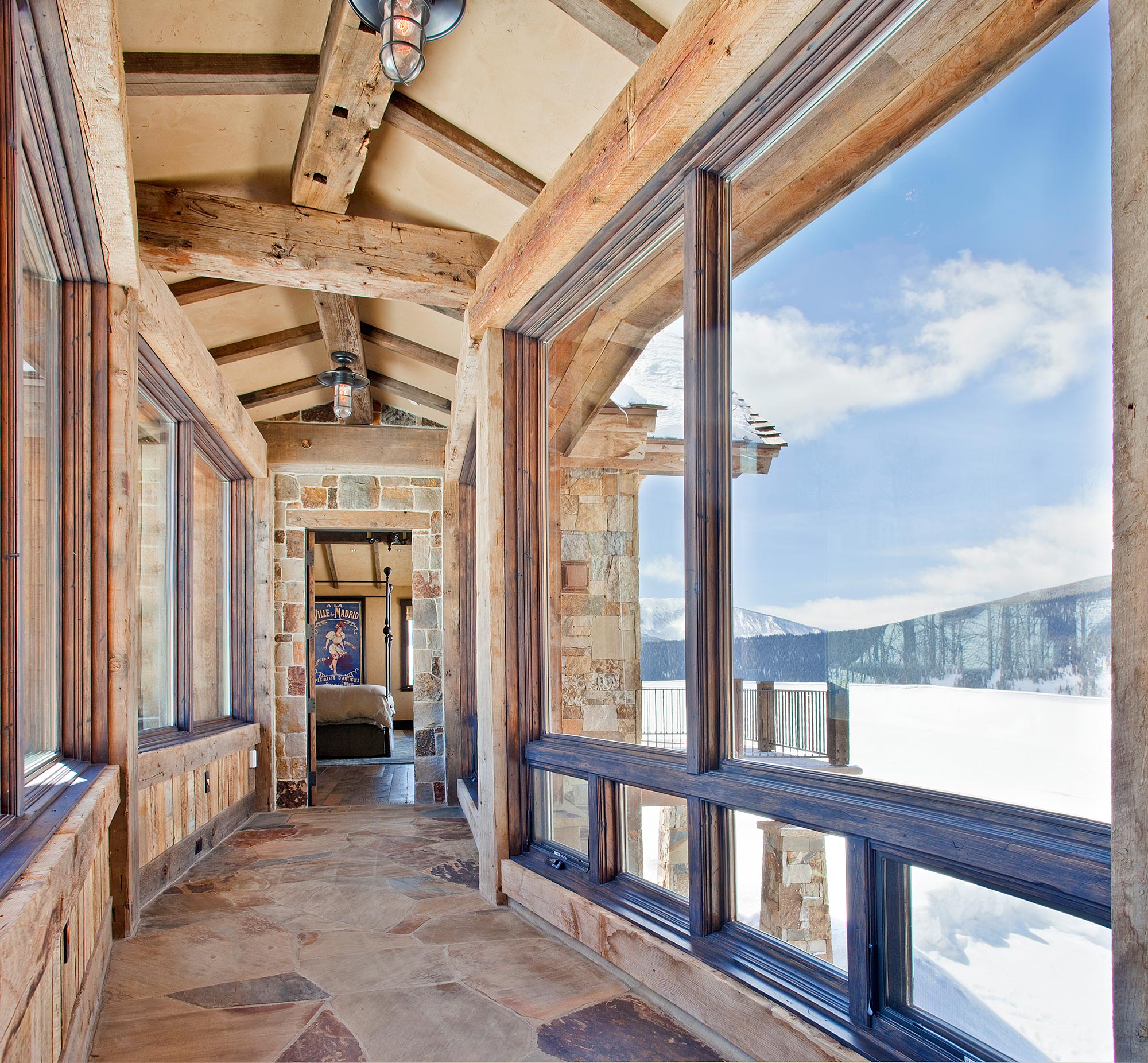 Glass corridor with reclaimed beams and flagstone floors framing snowy peaks