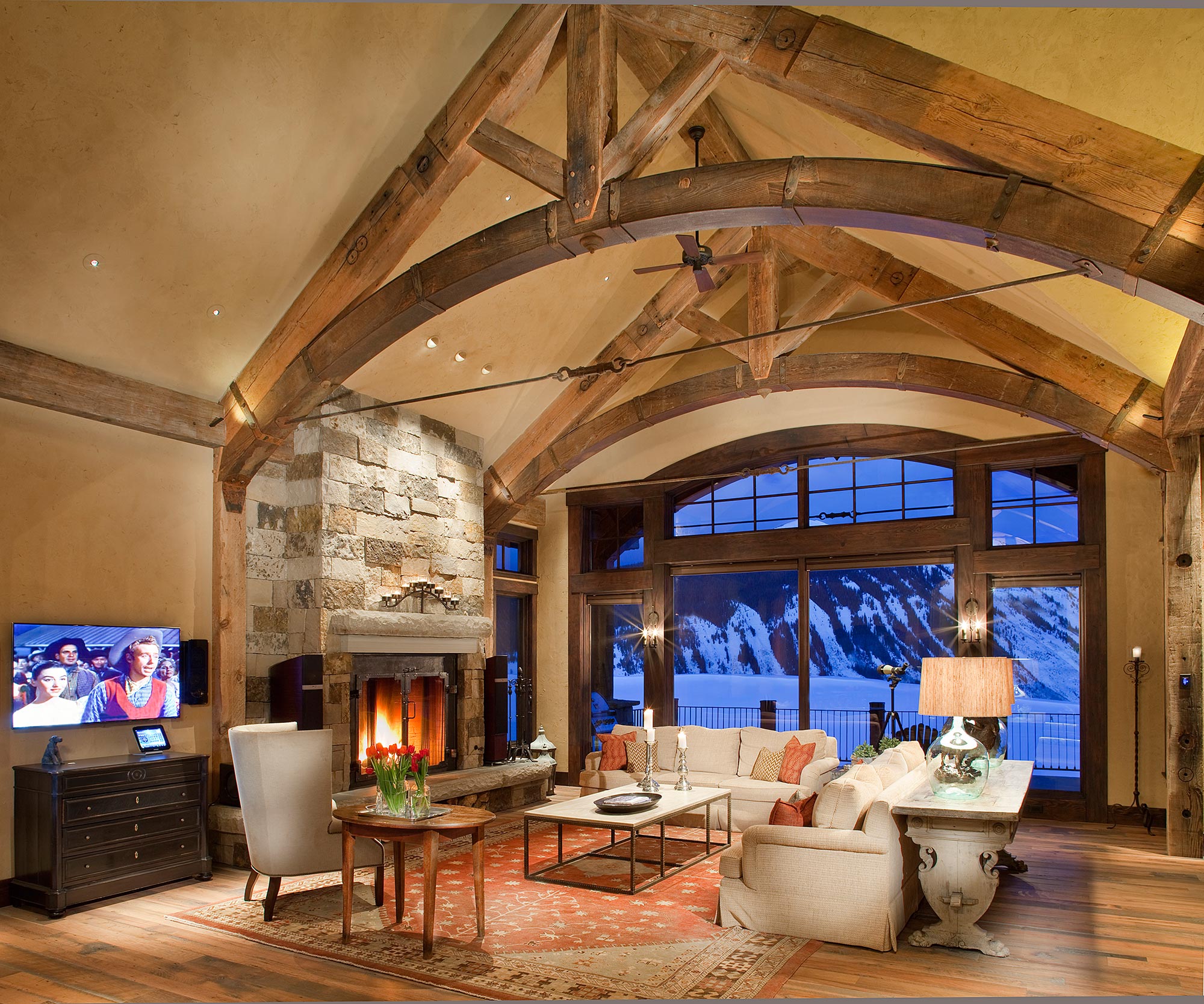 Great room with arched timber trusses and a stone fireplace at dusk